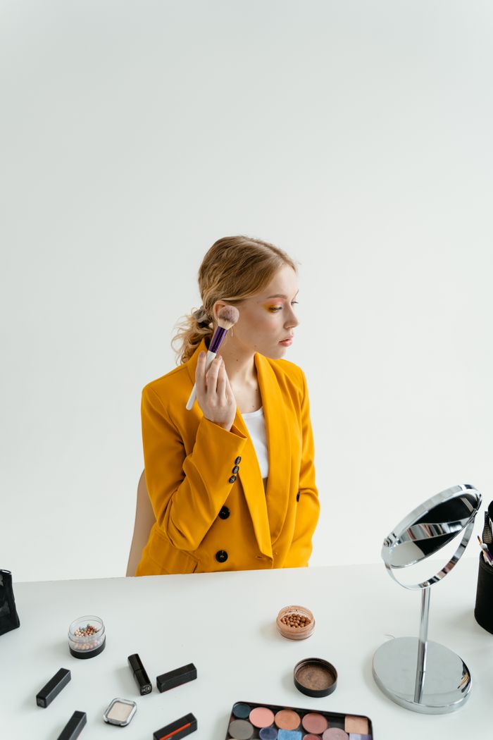 Woman in yellow blazer applying makeup using a brush at a vanity in a studio setting.